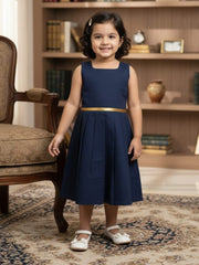 Young girl in a navy dress with a gold belt standing in a room with a bookshelf and chair.