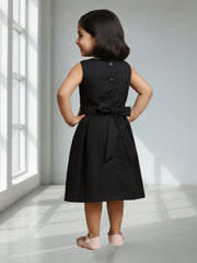 Young girl wearing a black dress with buttons and a bow, standing in a bright room.