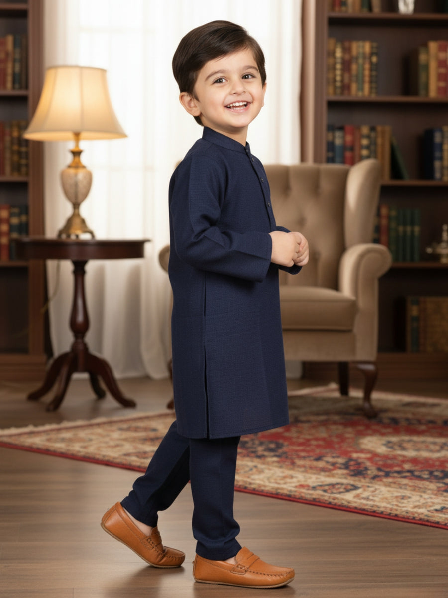Young boy in a navy kurta standing in a room with bookshelves and a lamp.