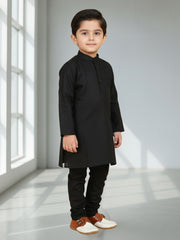 Young boy wearing a black kurta with white shoes in a bright room.