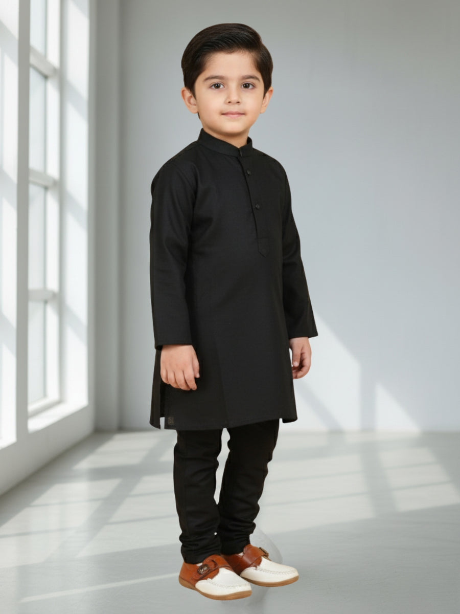 Young boy wearing a black kurta with white shoes in a bright room.