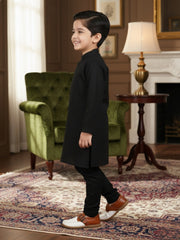 Young boy in a formal outfit standing in a room with a green armchair and lamp.